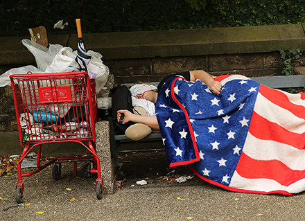 A homeless man sleeps under an American flag blanket on a park bench in New York City. New U.S. data reports a drop in the number of homeless people -- but not in New York and other states.